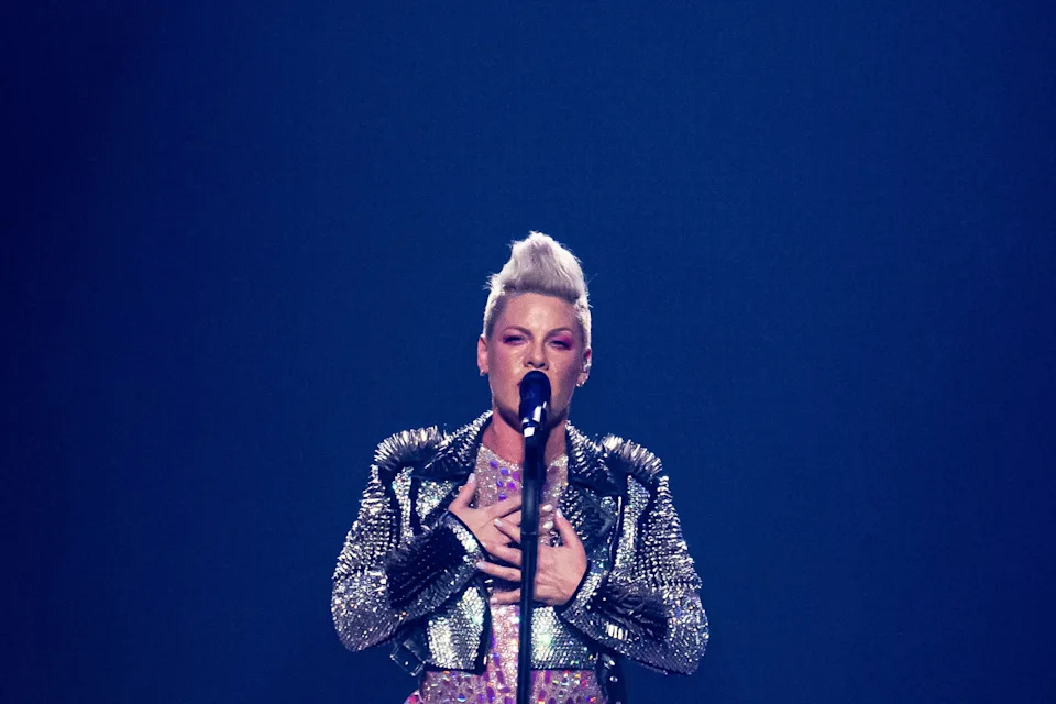 Pink performing on stage, wearing a sparkly studded jacket and patterned top, singing into a microphone with a focused expression