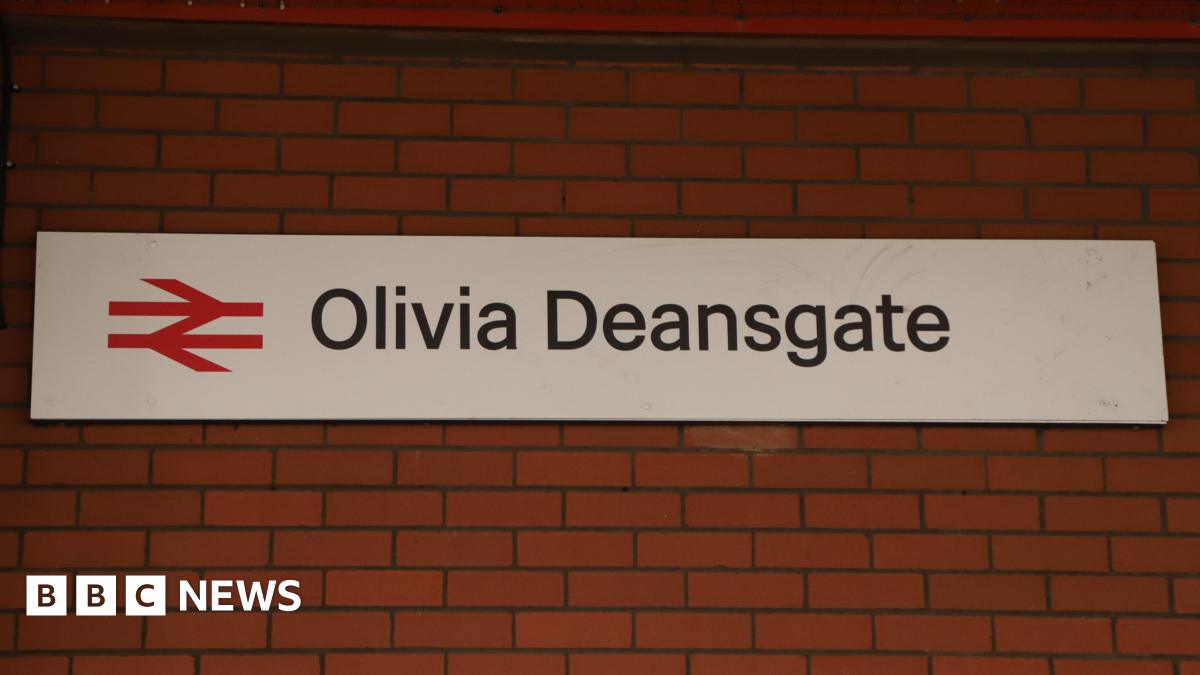 A train station sign reading 'Olivia Deansgate' hangs on a red brick station wall