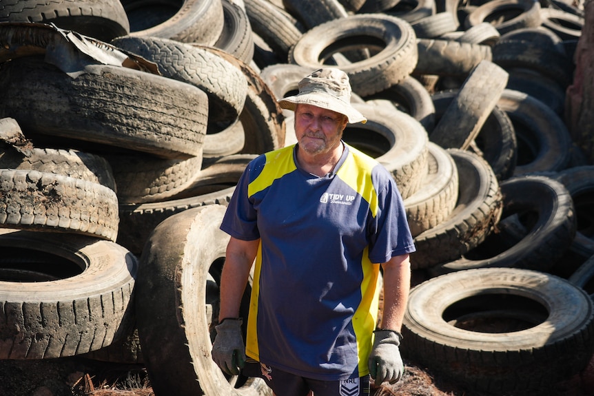 Man in blue and yellow shirt stands in front of waste tyres