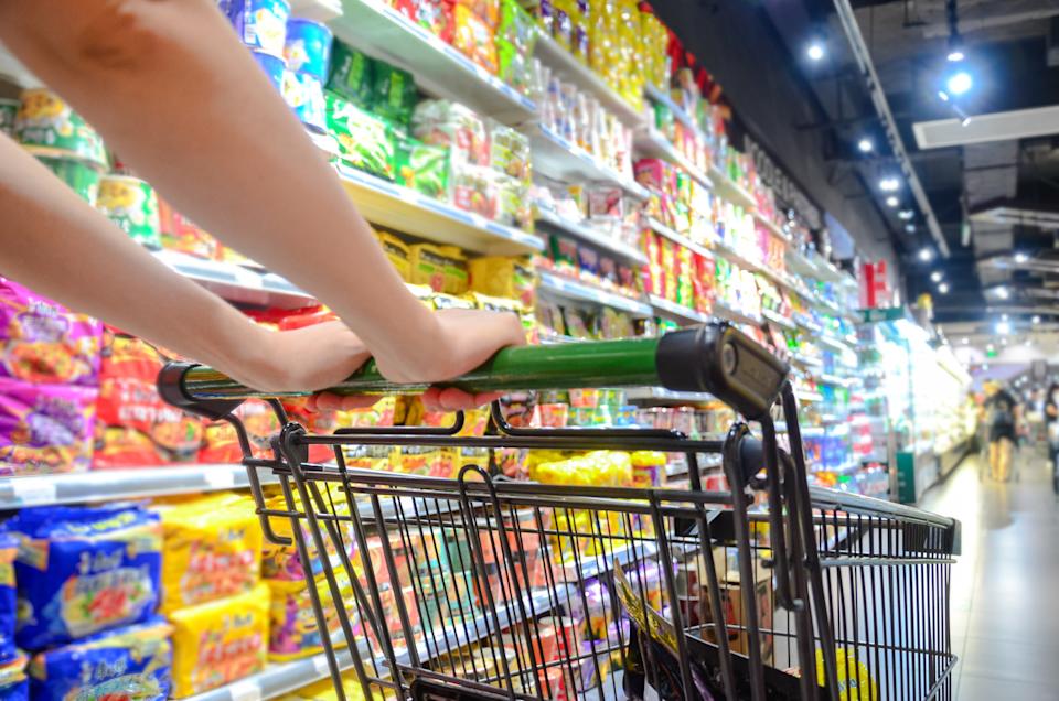 Person pushing a shopping cart down a supermarket aisle filled with various packaged goods neatly stacked on shelves