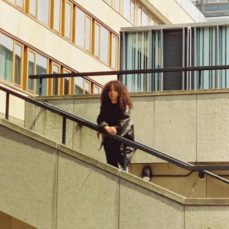 A person with long curly hair and a black jacket leans on a slanted handrail of a concrete outdoor staircase, with modern building windows in the background.