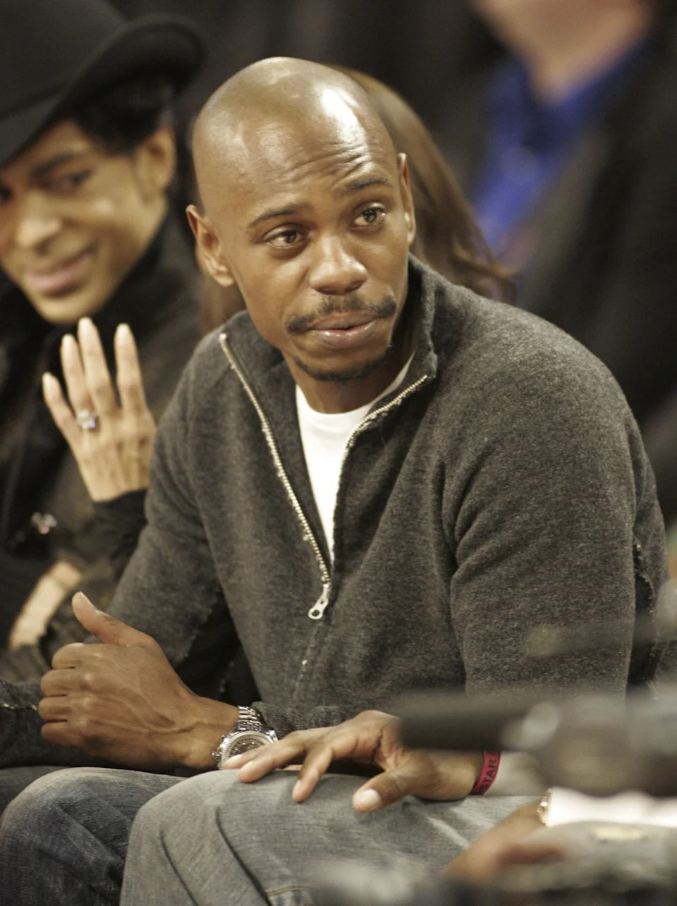 Two individuals sitting side by side in a lively setting, engaging in conversation. One is wearing a stylish hat, and the other a casual zip-up sweater