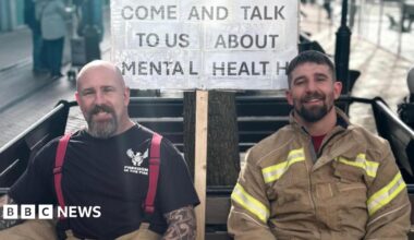 Firefighters Lee Ralph and Warren Shepherd dressed in uniform, sat on a bench. Behind them is a sign that says ' Come and talk to us about mental health'.