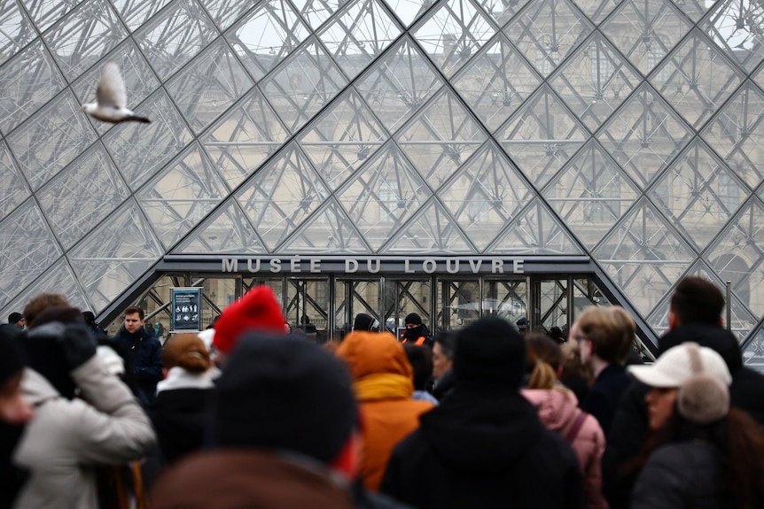 Queues of tourists in winter jackets standing in a group in front of the entry doors of the 'Musee Du Louvre'