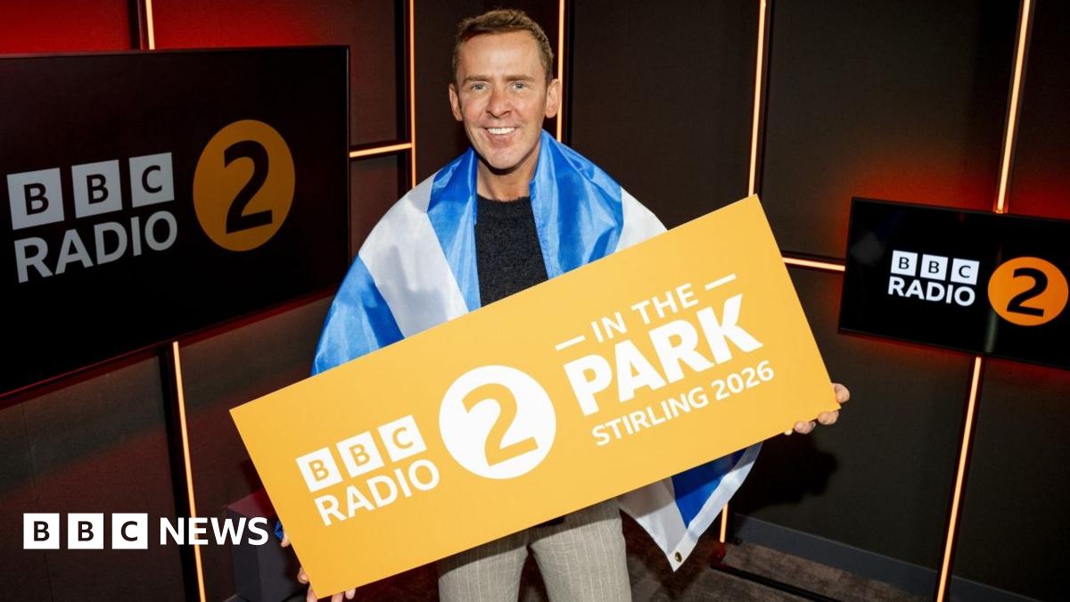 Scott Mills, wearing a Saltire flag and holding a Radio 2 In the Park Stirling 2026, in a radio studio