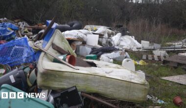 A large pile of fly‑tipped rubbish in a grassy area, including an old stained mattress, plastic crates, broken furniture and assorted household waste.