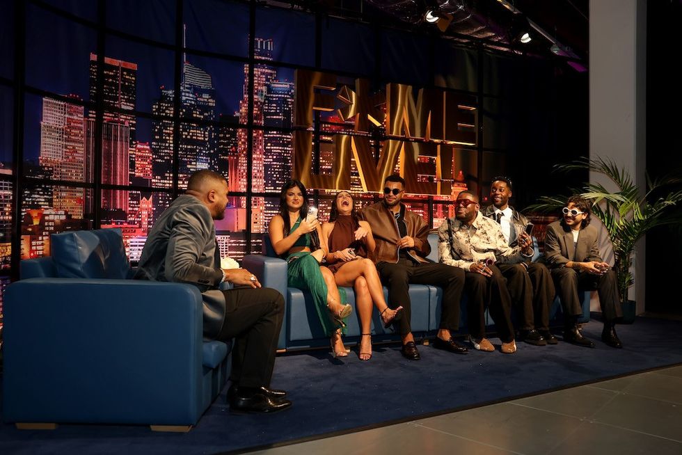 Fame Factory experience set - Talk show host interviews six stylish guests on a set with cityscape backdrop.