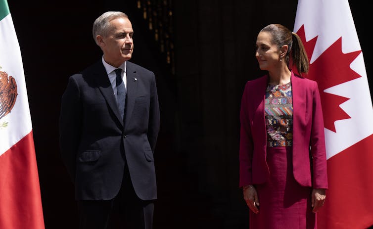 A woman in a pink suit and a man in a dark suit stand next to each other in front of the flags of mexico and canada