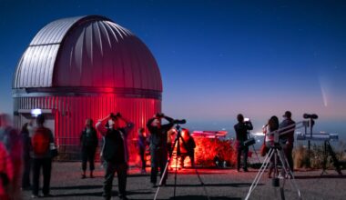 A circular dome of a telescope is illuminated in red light as a crowd of people holding binoculars and telescopes look at a pink and blue night sky.