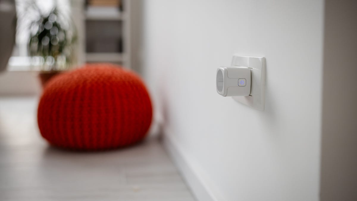 A smart plug plugged into a white home wall in front of an orange cushion.