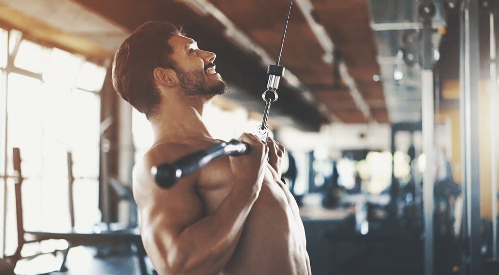 closeup side view of a muscular mid 20s shirtless man doing a back workout with a cable pull down machine. hes doing reverse grip pull downs with a visible pain grimace on his face. blurry gym equipment in background.