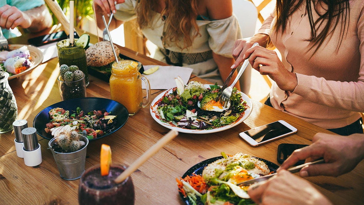 Young friends having fun eating brunch at healthy food restaurant, salads, veggie burgers, fried eggs and smoothies and juices seen on table.