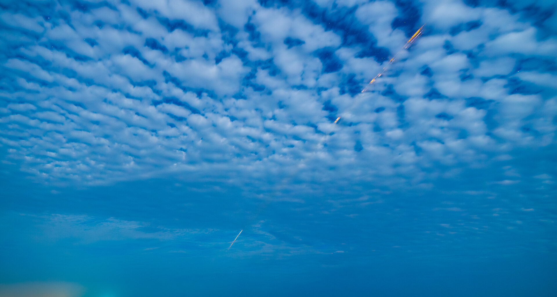 An orange and white streak cuts diagonally through a cloudy sky and down to Earth in the distance. The clouds have an unusual, rippling, wave-like pattern.