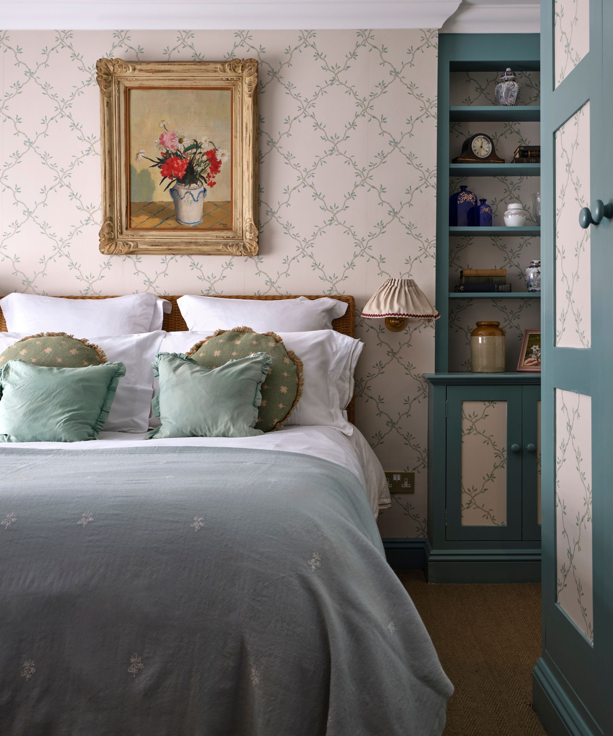 A bed with light blue linens and green decorative pillows set against a delicate floral trellis wallpaper. A teal built-in bookshelf sits to the right, filled with blue and white ceramics.