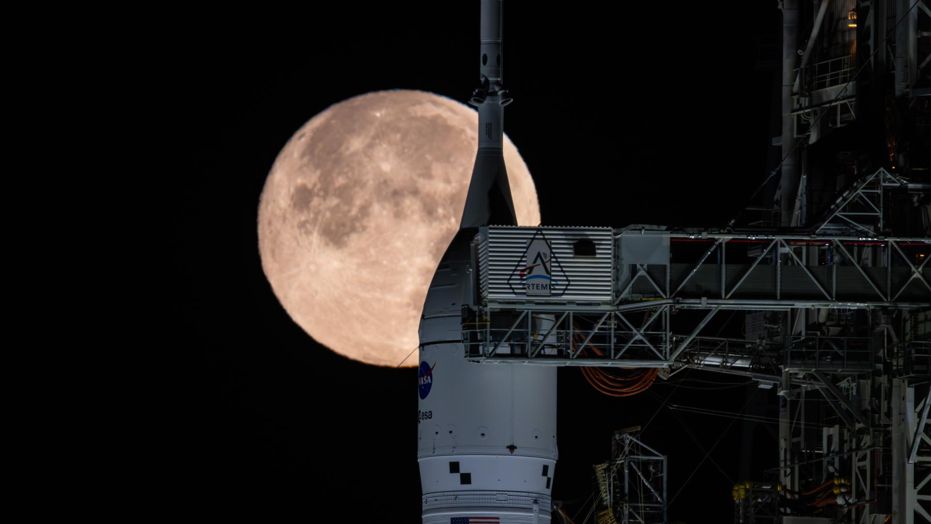A white rocket nose with the circular NASA logo stands next to the metal gantry underneath a glowing large full moon in a black night sky