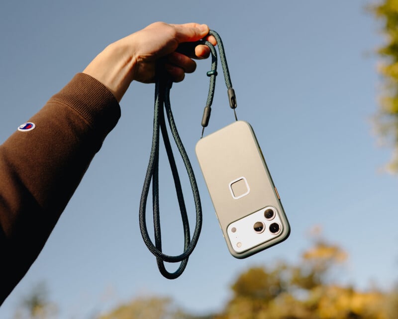 A hand holds up a smartphone attached to a green lanyard against a blue sky background, with trees blurred in the distance. The phone has a case with a square design near the center and three camera lenses visible.