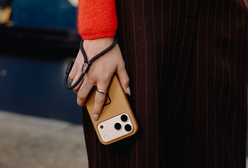 A person wearing a red sweater and brown pinstripe pants holds a beige smartphone with a black hand strap, showing a gold ring on their finger. The background is blurred.