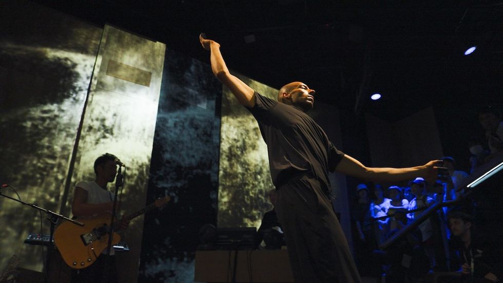 Performer on stage with arms outstretched, guitar player in background, blue lighting.