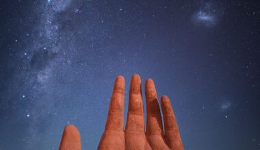 A large, surreal sculpture of a hand rises from the desert, under a starry night sky illuminated by the Milky Way