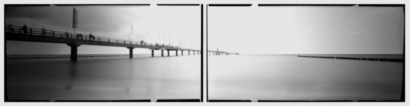 A black and white panoramic photo shows a long pier extending over calm water, with people walking along the pier. The sky is bright, and the horizon is visible in the distance. The image has a soft, surreal quality.