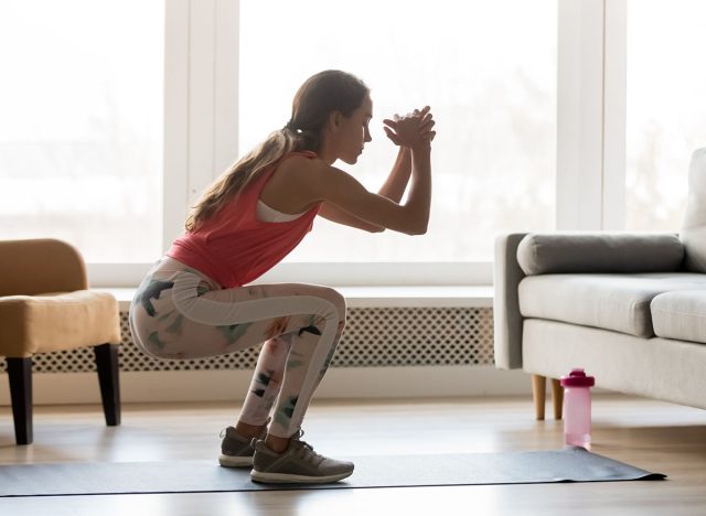 Sporty young woman doing squat morning exercise alone in living room, serious fit girl wearing sportswear crouching training muscles workout at home for healthy body lifestyle concept, side view