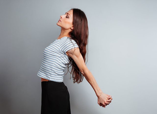 Sporty beautiful woman in casual clothing doing the chest and shoulders stretching exercise holding the palms in fist lock behind the back to relaxing body on blue studio background. Health lifestyle