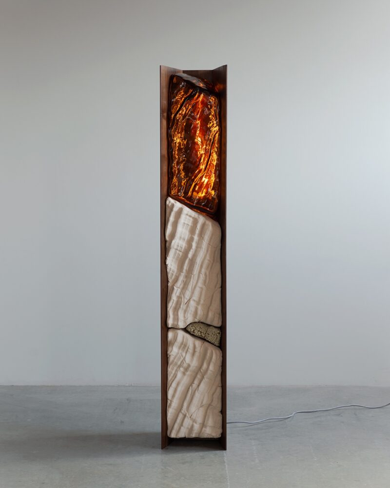 Tall sculptural floor lamp with a wooden frame, featuring illuminated amber-like resin and white stone-like segments, standing against a plain wall with a visible power cord.