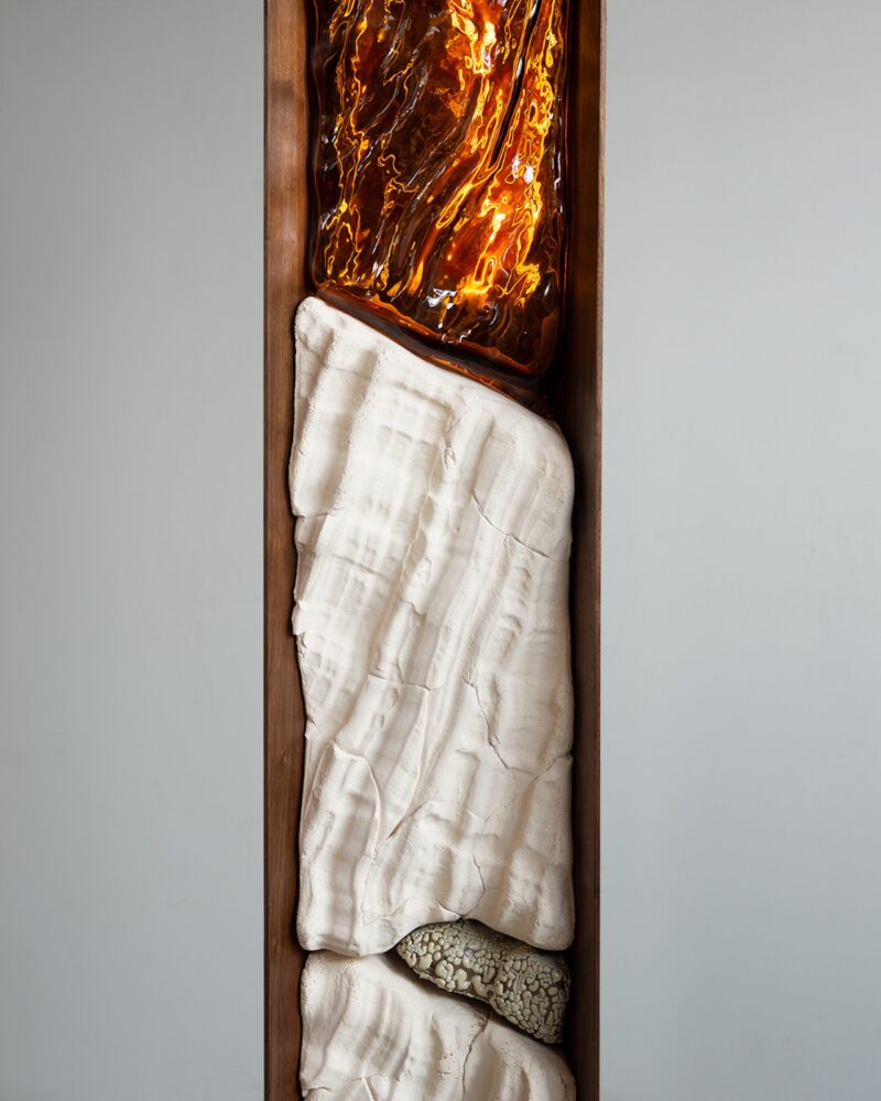 Vertical sculpture with sections of textured white material, rough beige stone, and a glossy, orange glass-like top, all encased in a rectangular wooden frame against a plain background.