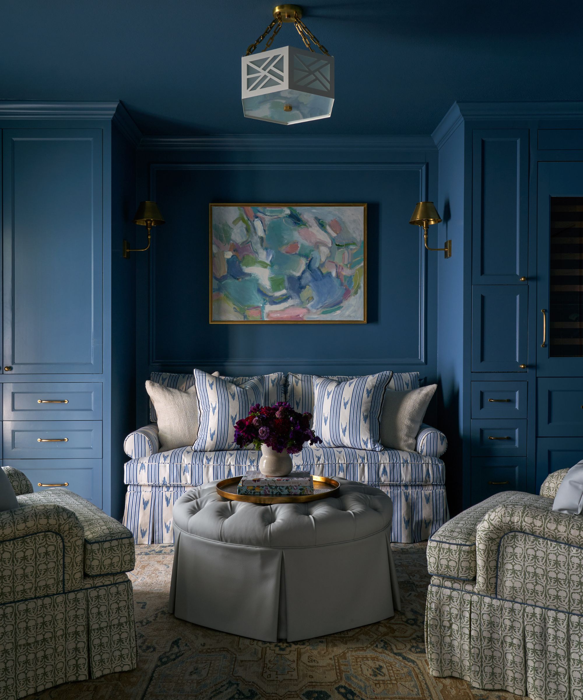 A living room with dark blue color-drenched walls, a blue and white sofa in the alcove with matching cushions, a gray pouffe, and two gray patterned armchairs.