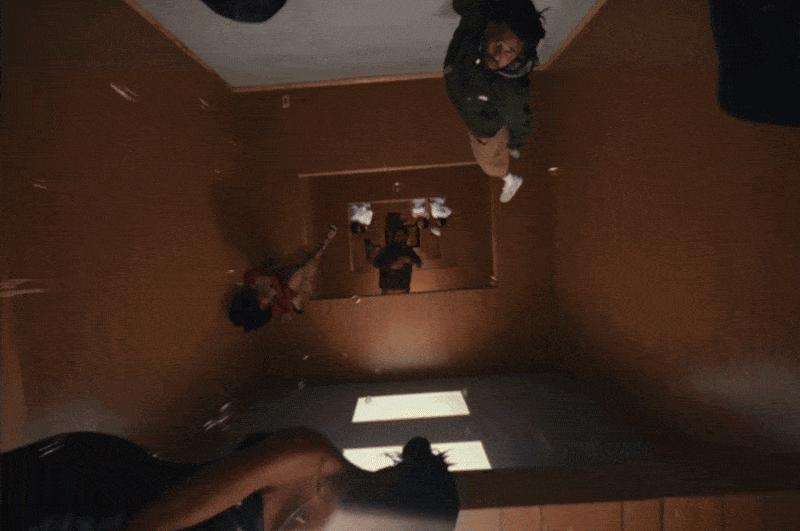a gif from a J. Cole music video directed by Ryan Doubiago, depicting a moving shot looping through different eras of the rapper's life, all taking place in the same studio walls. in this shot, a cork and drops of champagne float through the air in a party setting.