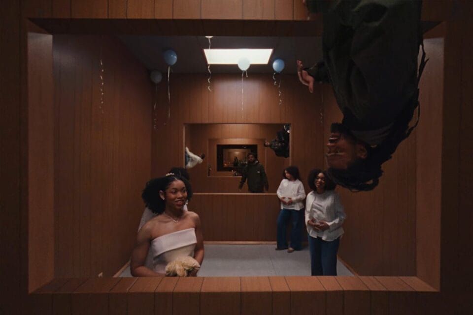 a still from a J. Cole music video directed by Ryan Doubiago, depicting a moving shot looping through different eras of the rapper's life, all taking place in the same studio walls. in this shot, the artist is reminiscing marriage and a child.