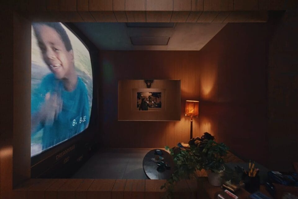 a still from a J. Cole music video directed by Ryan Doubiago, depicting a moving shot looping through different eras of the rapper's life, all taking place in the same studio walls. in this shot, the musician is reminiscing a childhood home video,