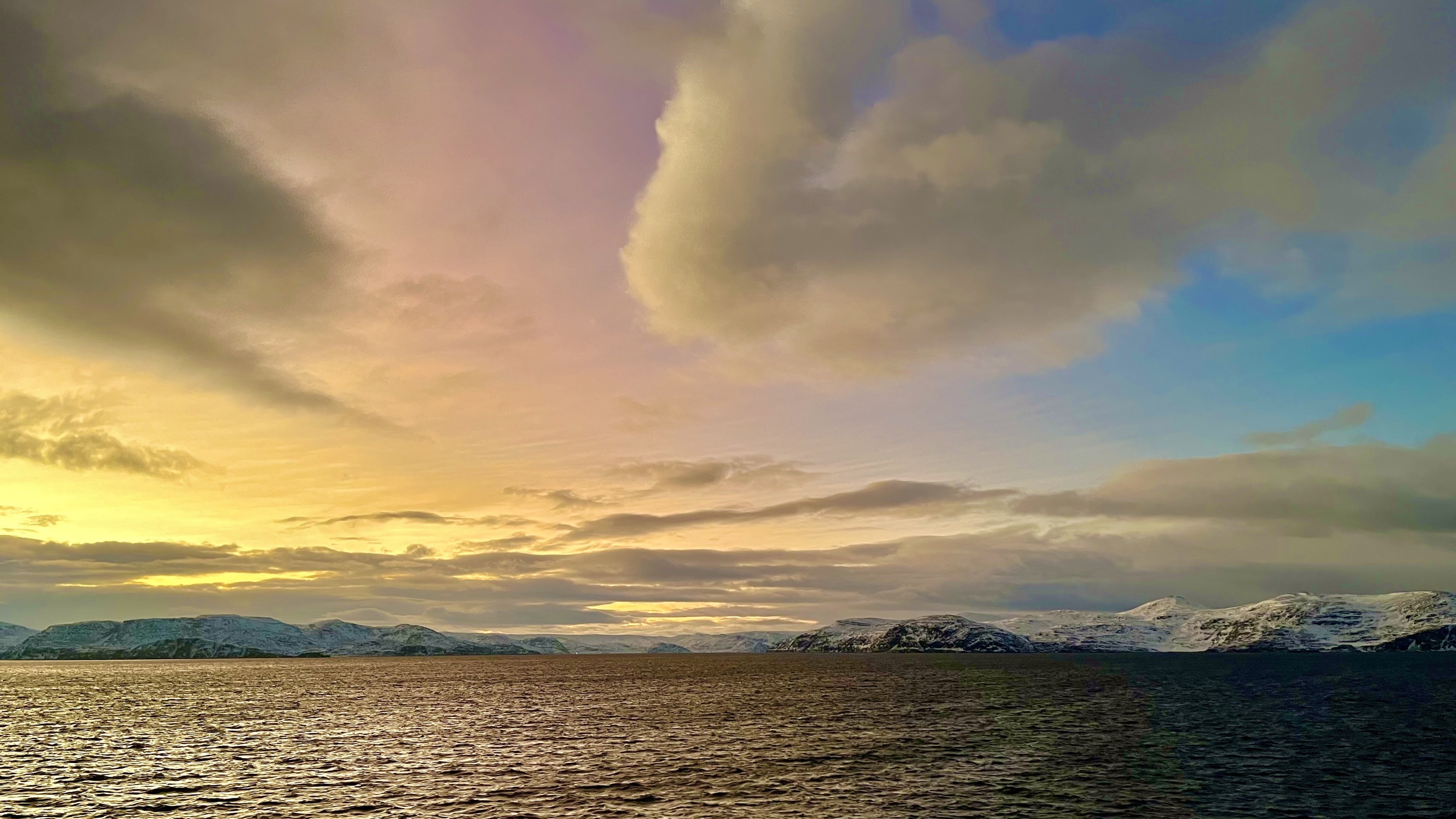 vibrant orange, pink and lilac sky above snow capped mountains and water below.