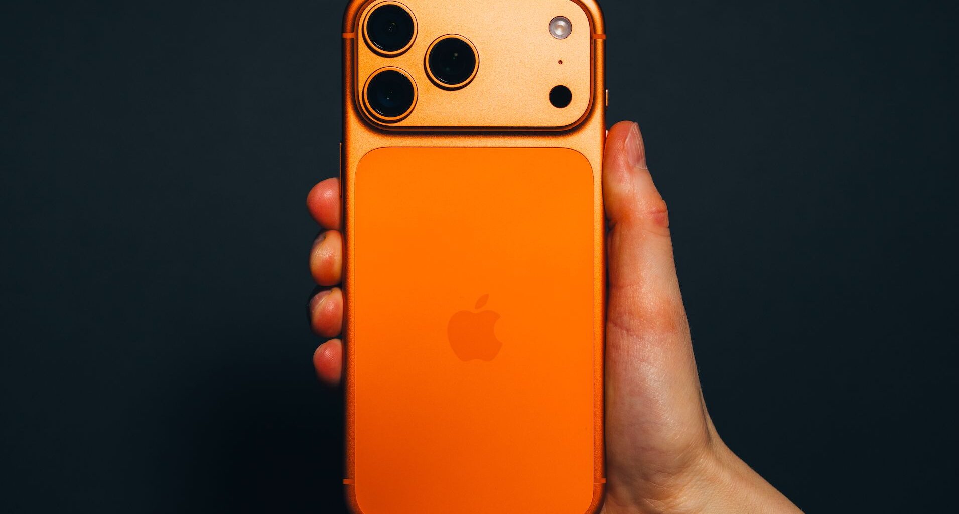 Krakow, Poland - November 6, 2025. Close-up editorial image shows a hand holding the latest Apple iPhone 17 Pro Max in Cosmic Orange, against a black background, highlighting the phone&rsquo;s unique colour and camera design. The shot captures current consumer technology trends and the distinct design choices of Apple devices. This editorial photo is suitable for articles, reviews, and technology news related to smartphones, Apple products, or mobile innovation.