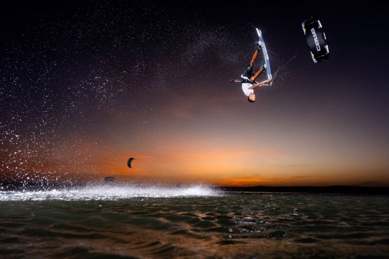 A person kiteboarding performs an aerial trick above the water at sunset, with water spray and another kiteboarder visible in the background.