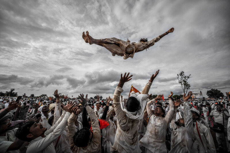 A group of people dressed in white joyfully toss a person high into the air during a lively outdoor celebration, with dramatic clouds filling the sky above the large gathered crowd.