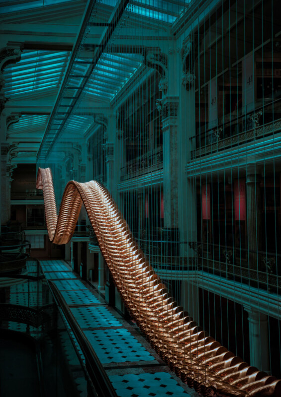 A large, illuminated, wave-shaped sculpture hangs in the atrium of a grand building with tall columns, glass ceilings, and patterned floors under teal lighting.