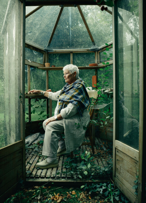 An elderly woman with short white hair sits on a wooden chair inside a small, glass greenhouse, surrounded by plants. She wears light clothing and a blue-green plaid shawl, looking outside through an open door.