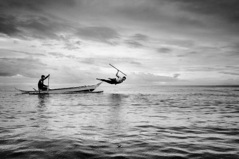 A black and white photo of a person paddling a small boat on the water, while another person leaps mid-air into the sea holding a stick, with clouds and horizon in the background.