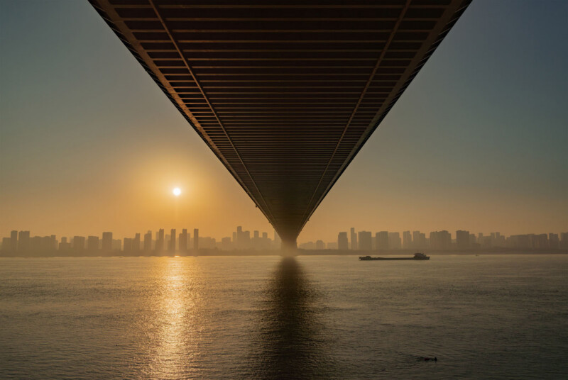 A view from beneath a large bridge, with the sun setting over a city skyline in the distance and its reflection shimmering on a calm river below. A boat is visible on the water.