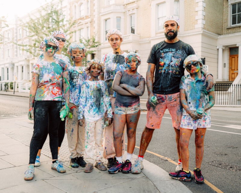 A group of seven people, including adults and children, stand on a city street covered in colorful paint splatters, looking at the camera. They wear goggles, shower caps, and casual clothes. Houses line the street behind them.