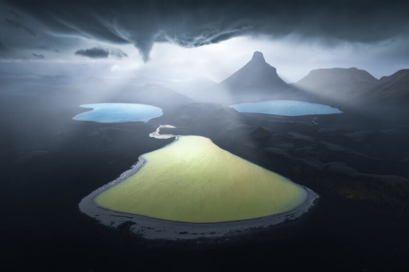 A dramatic landscape with three lakes—one greenish-yellow in the foreground and two blue in the background—surrounded by dark terrain, under stormy skies with sunlight breaking through, and a mountain peak in the distance.