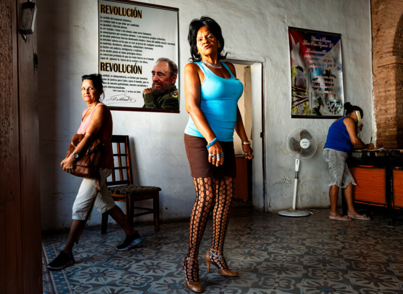 A woman in a blue top and patterned tights poses confidently indoors; another woman walks by, and a third works at a counter. Posters of Fidel Castro and revolutionary messages hang on the wall behind them.