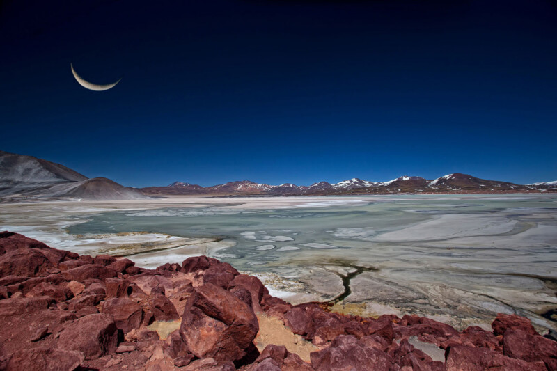 A crescent moon hangs in a deep blue sky over a barren, rocky desert landscape with red stones in the foreground and snow-capped mountains in the distance. Patches of salt flats and still water cover the ground.