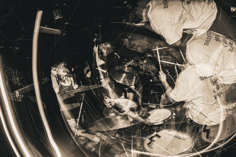 A high-energy, sepia-toned photo of a live punk band performance, with a vocalist passionately singing and musicians playing drums and guitar. Light trails and motion blur emphasize the chaotic atmosphere.