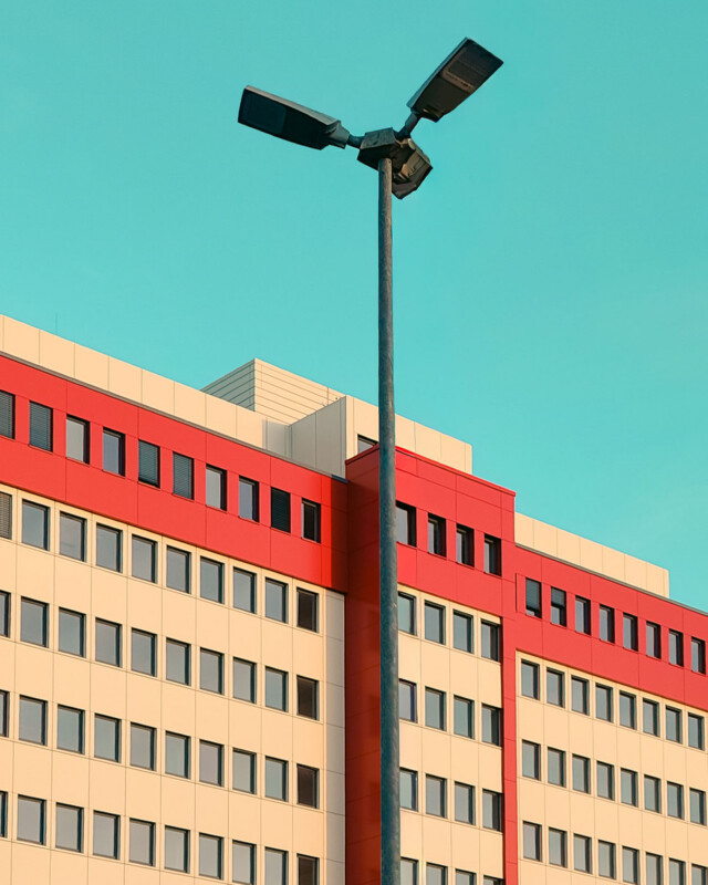A tall streetlight stands in front of a modern, multi-story building with red and beige horizontal stripes and rows of windows, set against a clear blue sky.