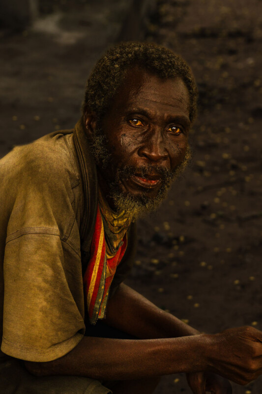 A man with curly hair, a beard, and mustache sits outdoors, looking into the camera. He wears a brown shirt and a striped scarf. The background is dark and blurred, highlighting his expressive face.