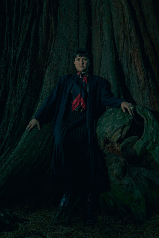 A person in a dark, flowing outfit and red scarf stands against the trunk of a massive tree, with one hand resting on a large twisted root in a dimly lit, forest environment.