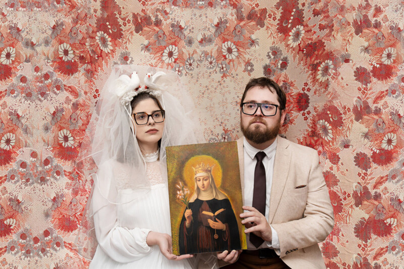 A solemn couple in vintage attire poses against a floral-patterned backdrop; the woman wears a white dress with a veil and decorative doves, while the man wears a light suit and glasses. They hold an icon of a crowned saint.