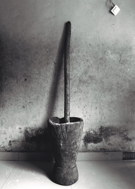 A traditional wooden mortar and pestle stands against a weathered, textured wall; an electrical socket is visible in the top right corner. The floor is tiled and the image is in black and white.